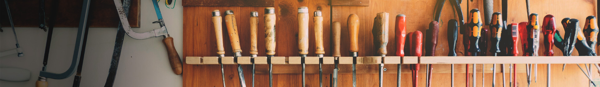 A wall with various hand tools neatly hung on a wooden shelf. The tools include wood handle chisels, screwdrivers, and a handsaw. The background is wooden, and the tools are in different colors and sizes