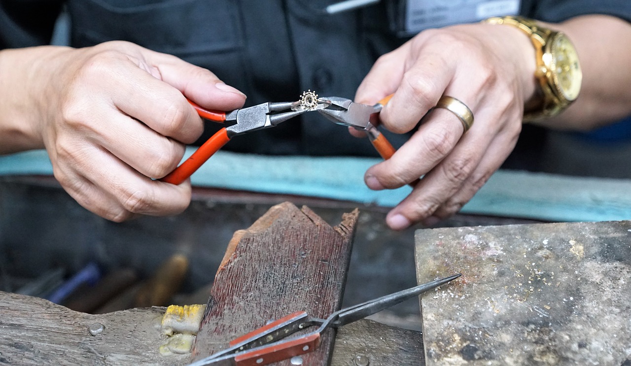 Twee handen van een vakman die met twee tangen een fijn sieraad bewerken. Op de werktafel liggen meer gereedschappen en een werkblad.