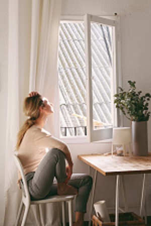 A woman sits on a chair next to a window that is open. She looks outside and has a thoughtful posture. The room is bright and friendly, with a table on which a plant stands - To the window handles category on tooler.eu/en